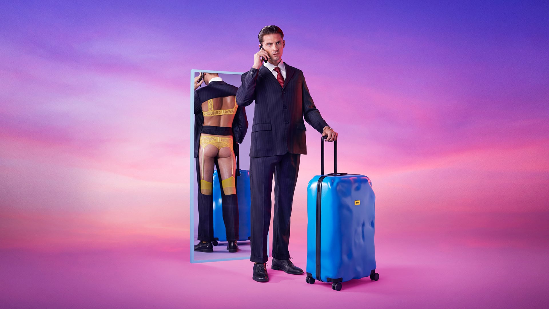 Campagna Crash Baggage: uomo in abito blu con trolley blu davanti a uno specchio che lo riflette in abiti colorati